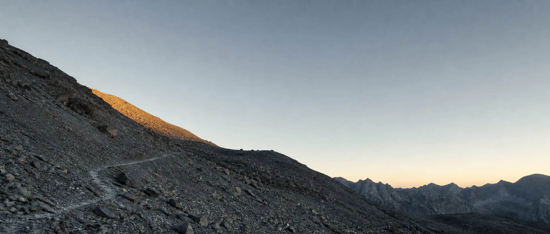 A scree slope at dawn, a faint switchback trail crossing the talus toward first light on the ridge